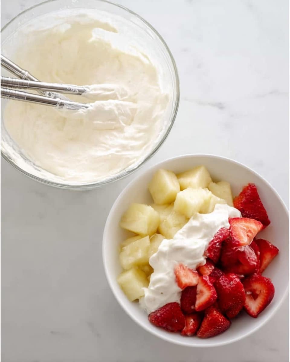 The image shows two parts: on the left side, a clear glass bowl with thick white cream inside and two metal beaters resting in the cream, placed on a white marbled surface; on the right side, a white bowl filled with three layers of cut fruit—light yellow pineapple pieces, light yellow banana slices, and red strawberry pieces—with a smooth white cream poured on top, sitting on a white marbled surface. photo taken with an iphone --ar 4:5 --v 7