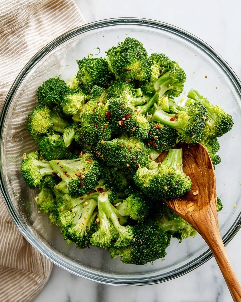 The image shows a white bowl filled with bright green roasted broccoli florets that have a slightly charred texture, giving a mix of dark and light green with some brown spots. On the edge of the bowl, there are three lemon wedges with a bright yellow rind and juicy pale yellow inside placed neatly. A silver fork is stuck into a broccoli piece, angled diagonally inward. The bowl sits on a white marbled surface, and the overall look is fresh and vibrant with a clean, simple presentation. photo taken with an iphone --ar 4:5 --v 7