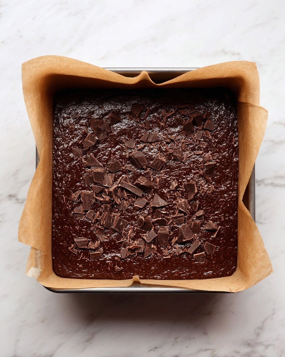 A baking pan lined with light brown parchment paper holds a thick, dark brown chocolate batter that fills the pan evenly. The batter has a shiny, slightly rough texture and is topped with small chunks and shavings of darker chocolate scattered across the surface. The pan sits on a white marbled surface. photo taken with an iphone --ar 4:5 --v 7