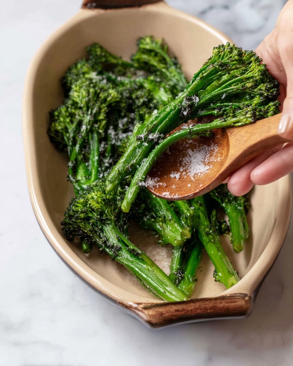 Inside a beige oval dish on a white marbled surface, bright green roasted broccolini with some charred edges are sprinkled with white salt crystals. A woman's hand is holding a small bunch of the broccolini over a smooth brown wooden spoon resting in the dish. The broccolini stems are long and firm with textured florets. photo taken with an iphone --ar 4:5 --v 7