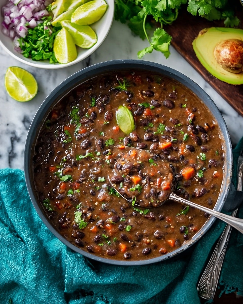 A close-up view of a dark grey pot filled with thick black bean stew showing black beans, diced carrots, and bits of green herbs mixed throughout the brownish bean soup. A silver spoon rests inside the pot, partially lifted with some stew dripping off. Two lime wedges and green cilantro leaves float on the stew surface near the top left. Behind the pot, a white bowl holds sliced avocado, chopped red onion, bright green cilantro sprigs, and lime wedges, all fresh and vibrant. Half an avocado with its brown seed sits on the white marbled surface to the right. A teal cloth is casually draped beneath the pot on the table. Photo taken with an iphone --ar 4:5 --v 7