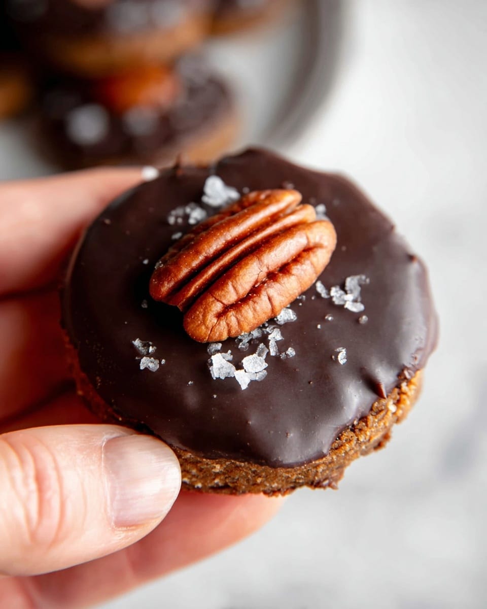 A close-up image shows a woman's hand holding a single cookie with three visible layers: the bottom layer is a rough-textured brown cookie, the middle layer is a thick, smooth, dark chocolate coating covering the entire cookie top, and the top layer is a shiny whole pecan nut placed at the center with a few flakes of coarse salt sprinkled around it, all held against a softly blurred white marbled texture background. photo taken with an iphone --ar 4:5 --v 7