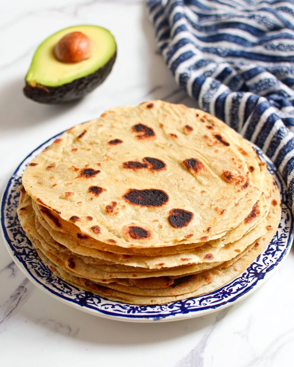 A stack of seven round flatbreads with a light brown color and dark brown charred spots lies on a white plate with blue ornate patterns at the edge. The flatbreads have a soft texture with uneven dark patches and slightly puffed areas, showing a warm and fresh look. Behind the plate, there is a halved avocado with a dark green outer skin, yellow-green flesh, and a large brown seed in the center, sitting on a white marbled surface. To the right, a folded blue and white striped cloth is partially visible, adding a cozy touch to the setup. photo taken with an iphone --ar 4:5 --v 7