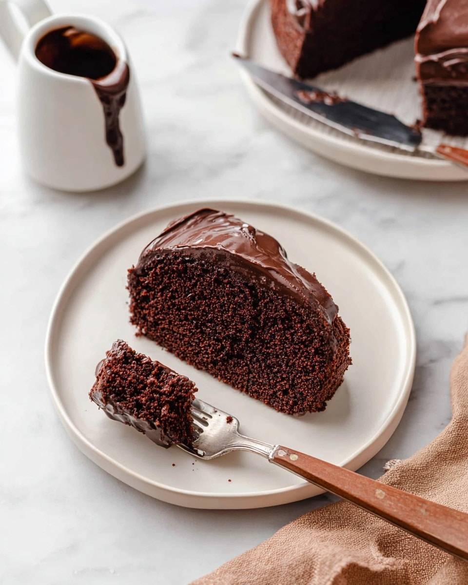 A single slice of dark chocolate cake with one smooth layer of shiny chocolate frosting on top sits on a white plate. The cake slice has a soft, crumbly texture with a deep brown color. A silver fork with a wooden handle holds a small bite of the cake near the slice. In the background, another white plate holds the rest of the cake with a knife that has some frosting on it. To the left is a small white pitcher filled with dark chocolate sauce dripping over the edge. Everything is placed on a white marbled surface with a light brown cloth partially visible at the bottom right corner. Photo taken with an iphone --ar 4:5 --v 7