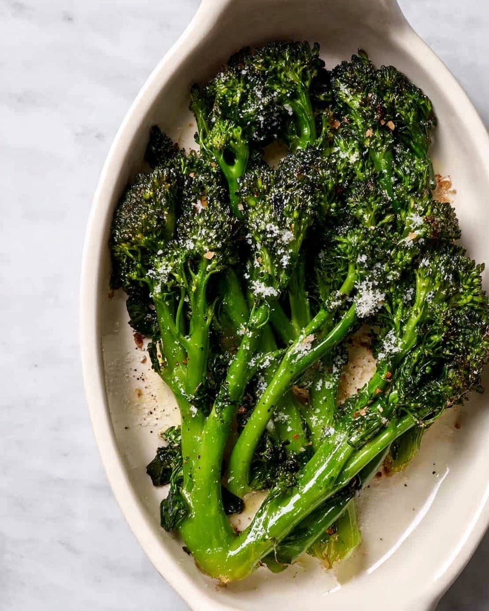 A white oval dish holds several pieces of roasted broccolini arranged loosely, with bright green stems and dark green, slightly charred florets. The broccolini is sprinkled with coarse white salt flakes, adding texture and contrast. Some parts of the stems and leaves have a slight crispiness from roasting, showing light browning. The dish is placed on a white marbled surface, giving a clean and fresh look. photo taken with an iphone --ar 4:5 --v 7