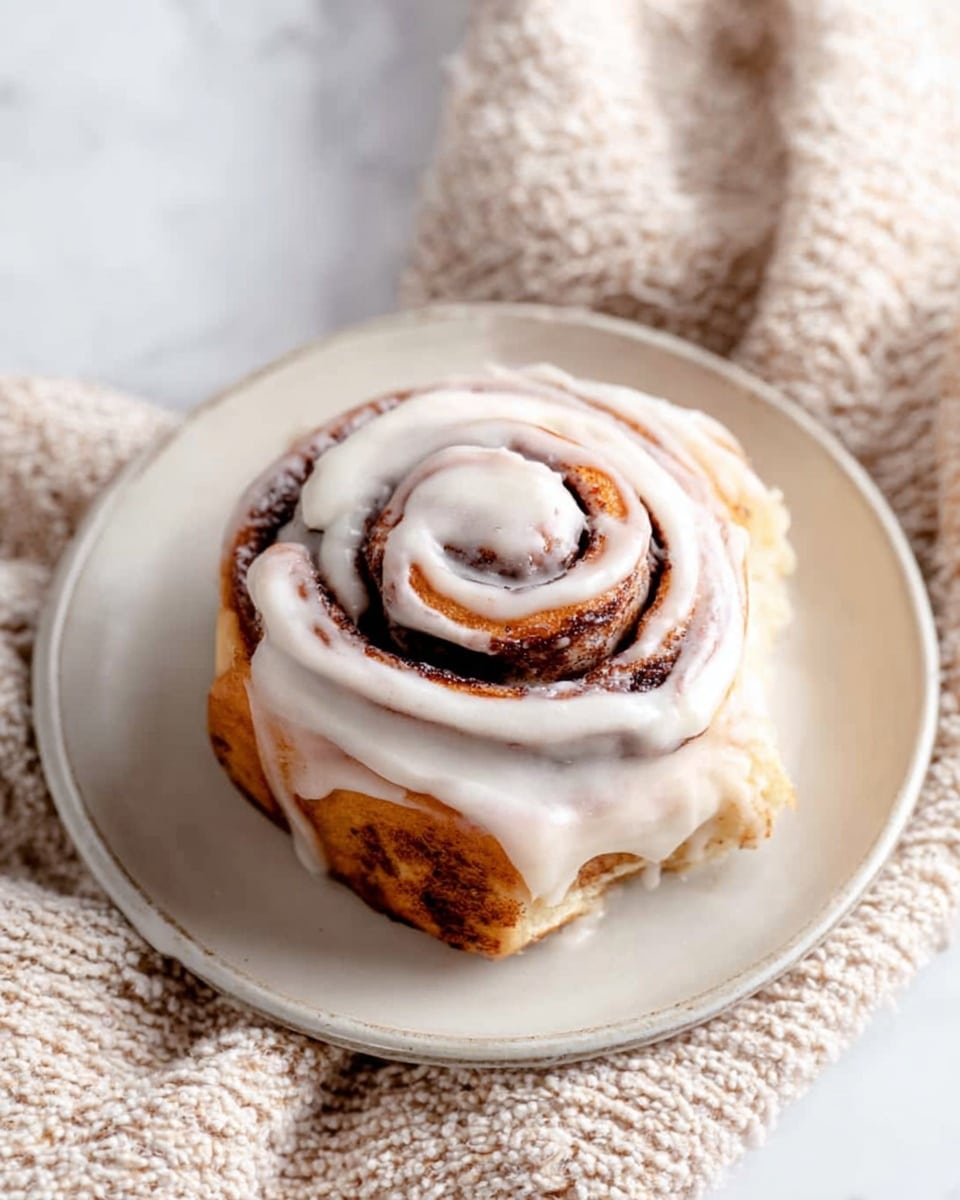 A single cinnamon roll sits in the center of a white round plate, placed on a soft, chunky beige cloth over a white marbled surface. The cinnamon roll has three visible spirals of golden-brown dough swirled tightly with dark brown cinnamon filling. A thick layer of white creamy icing is spread unevenly on top, mostly covering the roll but allowing some of the cinnamon swirl to show through. The icing has a smooth, shiny texture and softly drips down the sides of the roll. Photo taken with an iphone --ar 4:5 --v 7