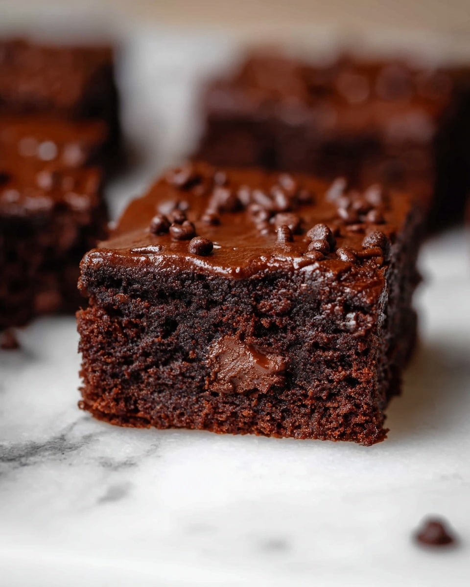 A close-up of a single square brownie piece with two visible layers; the bottom layer is thick and dense, dark brown with a moist and crumbly texture mixed with chocolate chunks inside, while the top layer is a smooth, glossy chocolate glaze sprinkled with small chocolate bits. The brownie rests on a white marbled surface, with blurred out chunks of brownies in the background. Photo taken with an iphone --ar 4:5 --v 7