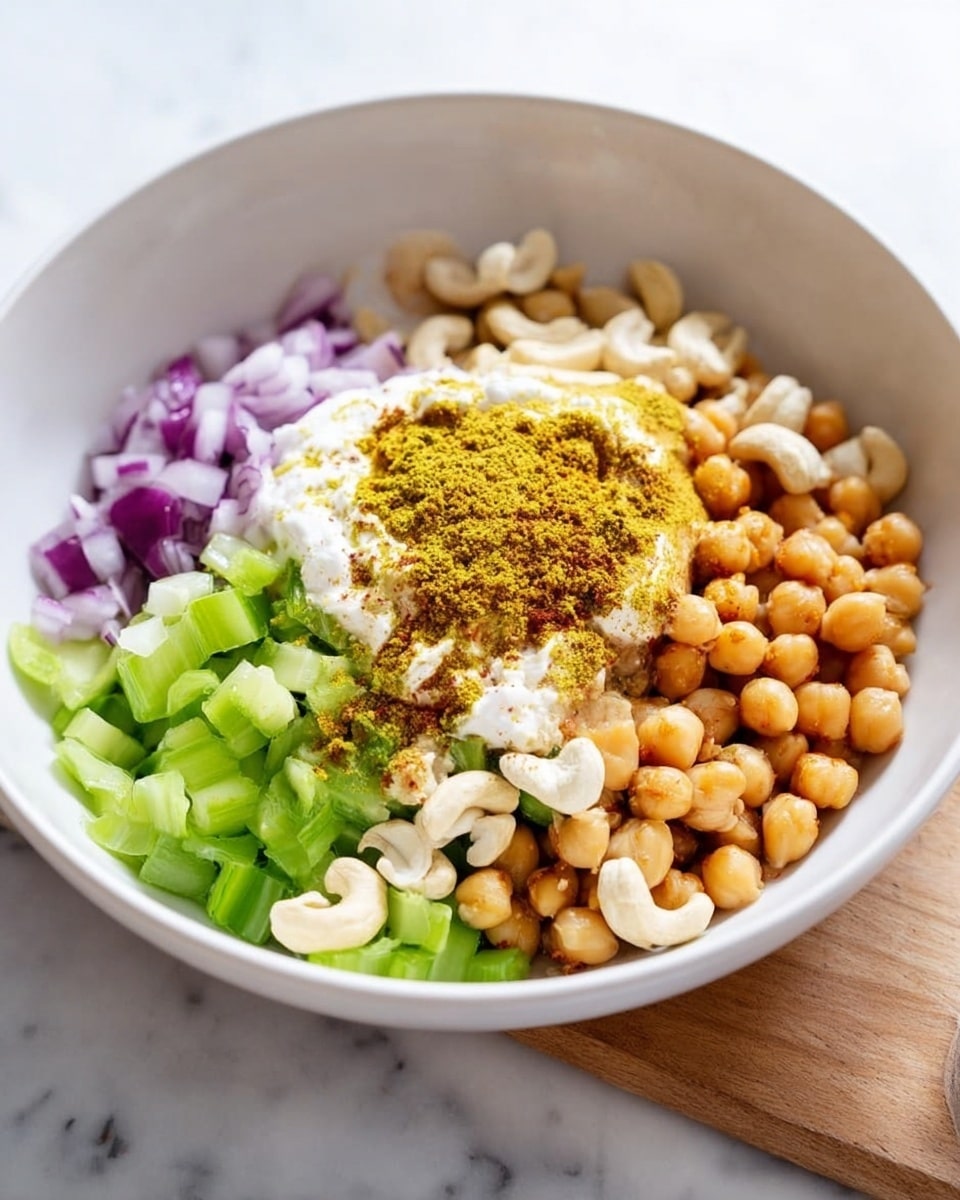 A large white bowl holds layers of ingredients: at the bottom are small pieces of purple onion on the left side; next to them are chopped green celery pieces forming a middle layer; beside the celery are pale yellow chickpeas clustered on the right side. On top of the chickpeas sits a dollop of thick white yogurt dusted with bright yellow curry powder. Scattered over the center are light beige cashew nuts. The bowl sits on a white marbled surface with a wooden board partly visible on the right side. Photo taken with an iphone --ar 4:5 --v 7