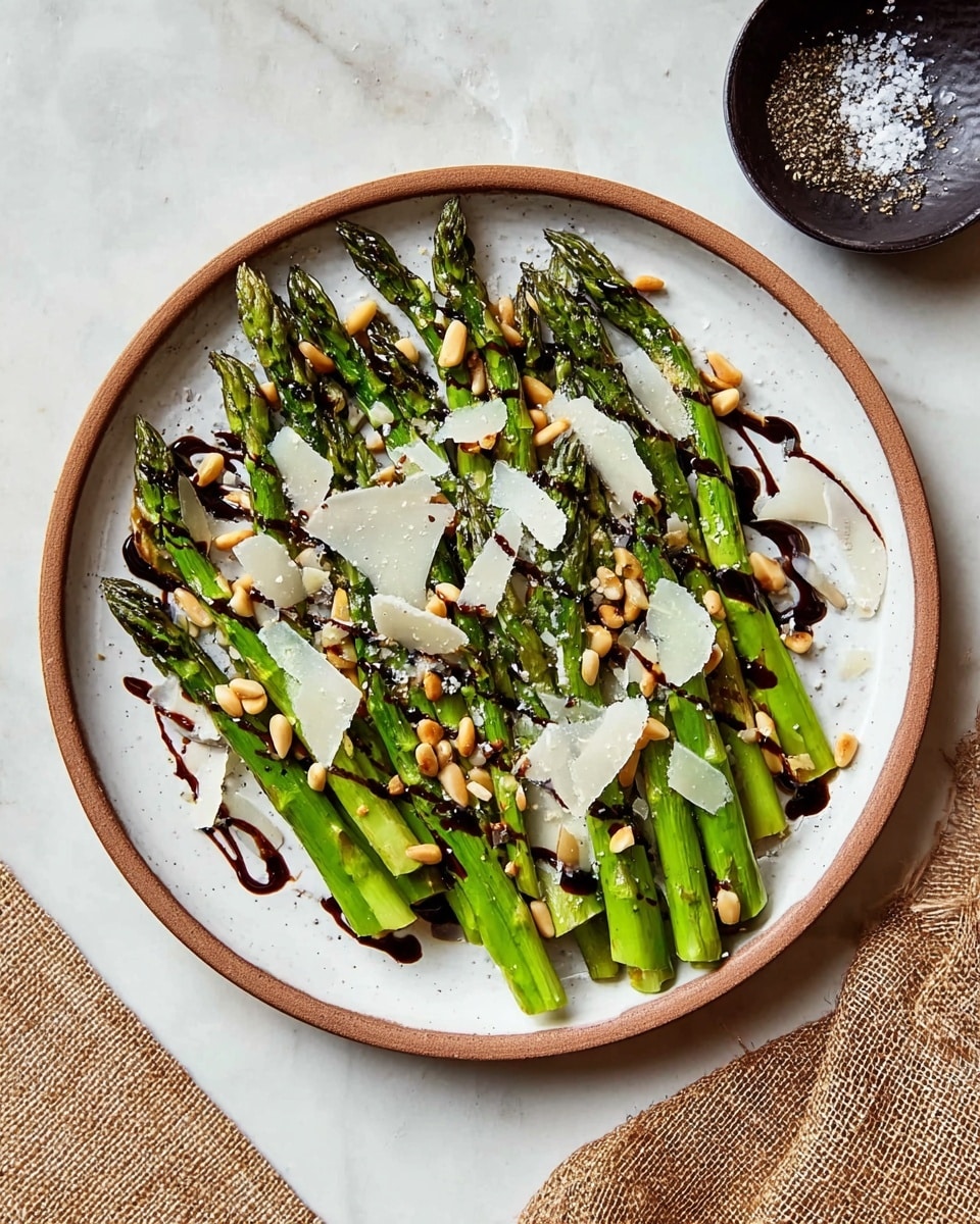 The image shows a white round plate filled with bright green grilled asparagus stalks arranged side by side, slightly overlapping, and spread across the plate. On top of the asparagus, there are thin, irregular white cheese shavings placed unevenly, some larger and some smaller. Small tan-colored pine nuts are scattered on the asparagus and cheese. The dish is drizzled with a dark balsamic glaze in thin lines running over the layers. The plate is set on a white marbled surface with a small dark round dish in the upper right corner containing coarse salt and cracked black pepper. The edge of a beige textured cloth is visible at the lower left corner. photo taken with an iphone --ar 4:5 --v 7
