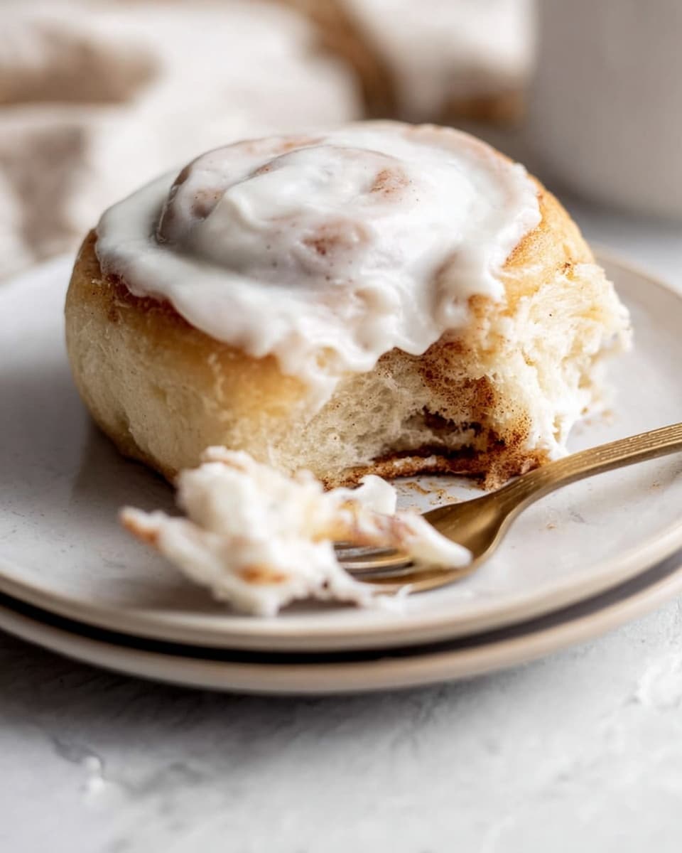 The image shows a cinnamon roll on a white plate with a white marbled texture background. The cinnamon roll has one main round layer with a light golden-brown color and a soft texture. On top, there is a thick layer of white icing that looks creamy and smooth. In front of the cinnamon roll, there is a piece torn off, revealing a slightly darker inner side with a brownish cinnamon swirl texture. A fork with white cream on its tines rests on the torn piece. The overall look is warm and soft with a mix of light browns and white colors. Photo taken with an iphone --ar 4:5 --v 7