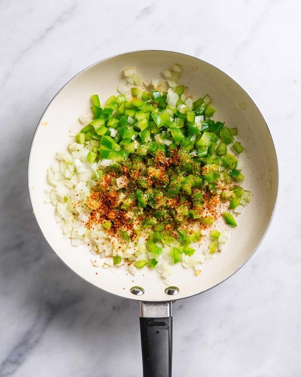 A white frying pan with a black handle is shown from above on a white marbled surface. Inside the pan, there is one layer of finely chopped white onions covered by a layer of bright green chopped bell peppers, lightly sprinkled with a layer of reddish-brown spices on top, creating a mix of colors and textures. The pan has three silver rivets where the handle meets the pan. photo taken with an iphone --ar 4:5 --v 7