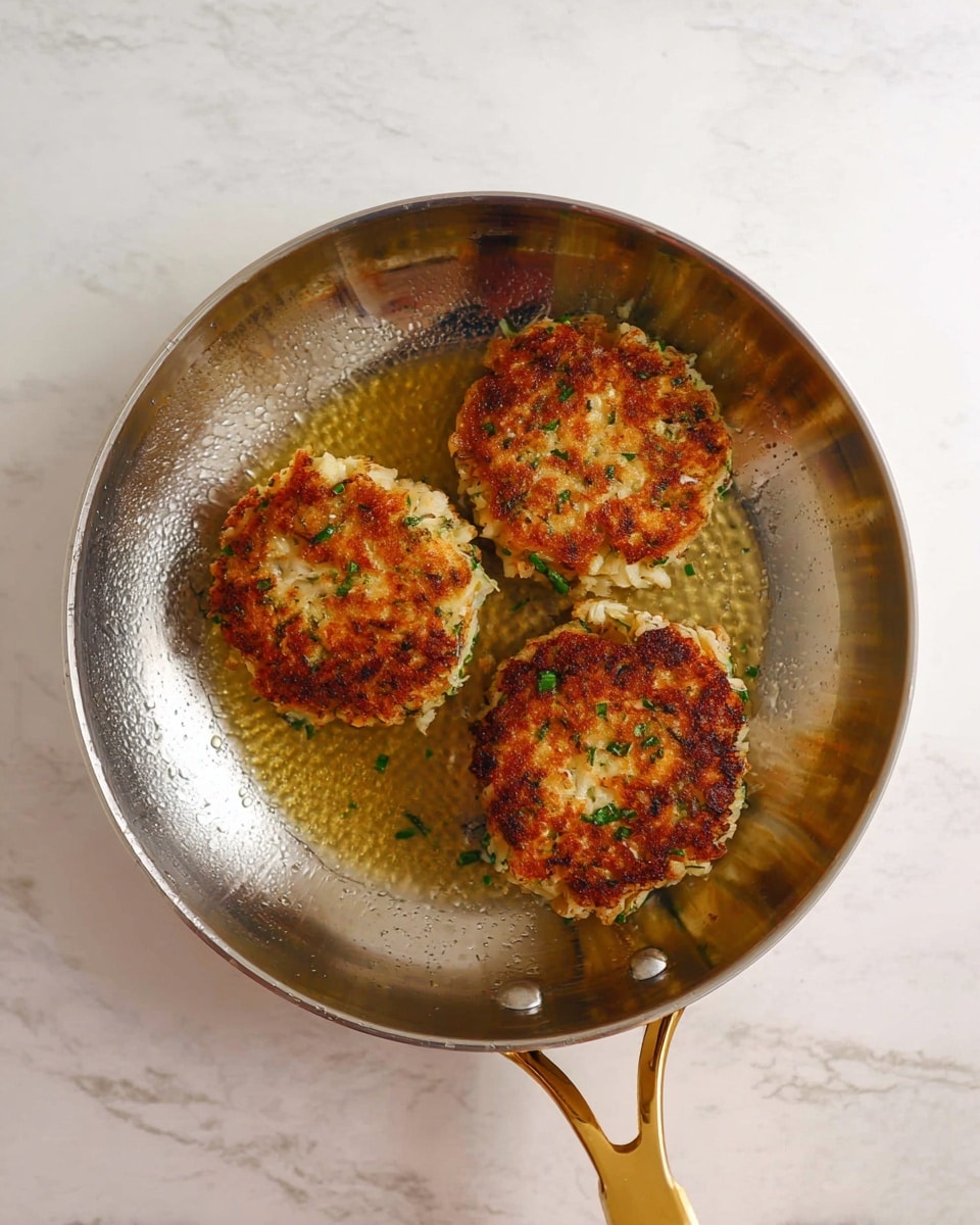 Three cooked crab cakes with a golden brown crust sit inside a shiny silver frying pan. The crab cakes show flaky white meat mixed with herbs and some finely chopped green bits. The pan is slightly oily around the crab cakes, indicating they were fried. The pan has a gold handle and rests on a white marbled surface. photo taken with an iphone --ar 4:5 --v 7