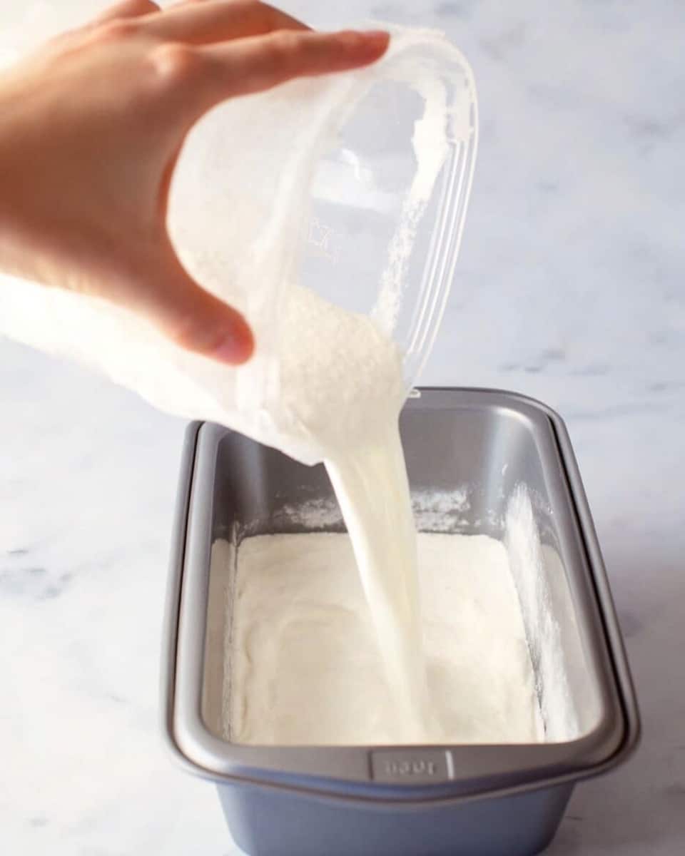 A woman's hand is holding a clear container pouring a thick white liquid into a rectangular metal bread pan. The liquid is smooth and creamy, filling the bottom of the pan with a light, even layer. The background surface is a white marbled texture. Photo taken with an iphone --ar 4:5 --v 7