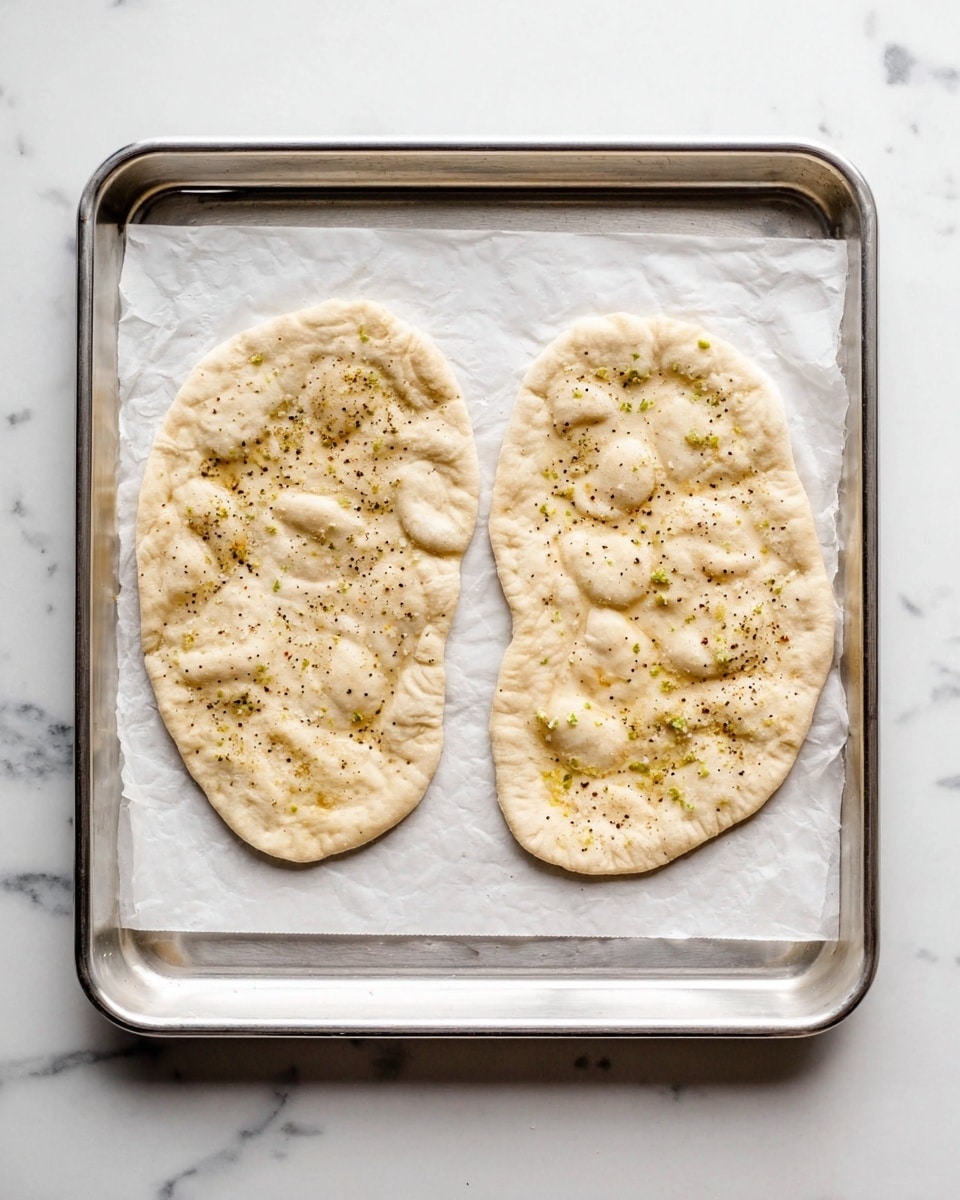 Two uncooked flatbreads placed side by side on a silver baking tray lined with white parchment paper. Each flatbread is irregularly shaped with a pale dough color and has visible small indentations and sprinkled black pepper across the surface. There are light greenish spots that suggest bits of seasoning mixed into the dough. The tray sits on a white marbled surface, adding a clean and bright background to the scene. photo taken with an iphone --ar 4:5 --v 7