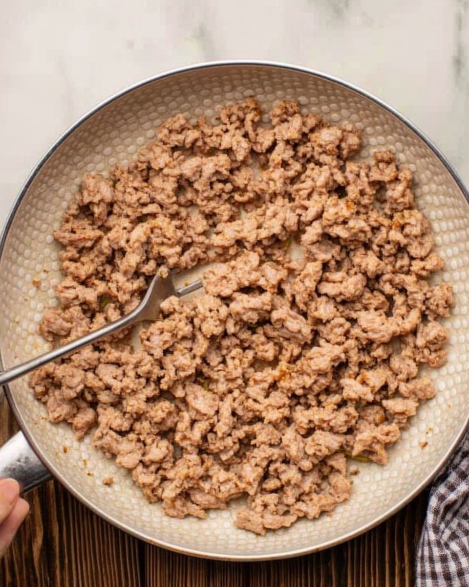 The image shows a white pan filled with many small pieces of cooked ground meat that are light brown in color, evenly spread across the pan's surface. The pan is placed on a white marbled surface, and part of a woman's hand holding a utensil is visible in the top left corner. The meat pieces look dry with some small bits of liquid in the center, and the pan has a textured bottom with a silver rim. photo taken with an iphone --ar 4:5 --v 7