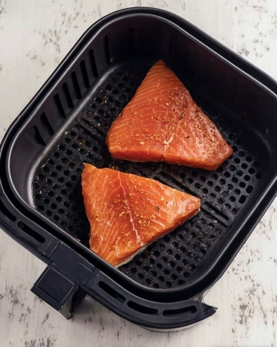The image shows two raw salmon fillets placed inside a black air fryer basket. The fillets are triangular in shape, with a rich orange-pink color and a slightly shiny texture due to a light coating of oil or marinade with some visible seasoning specks on top. The air fryer basket has a grid pattern at the bottom and is set on a white marbled surface. Photo taken with an iphone --ar 4:5 --v 7