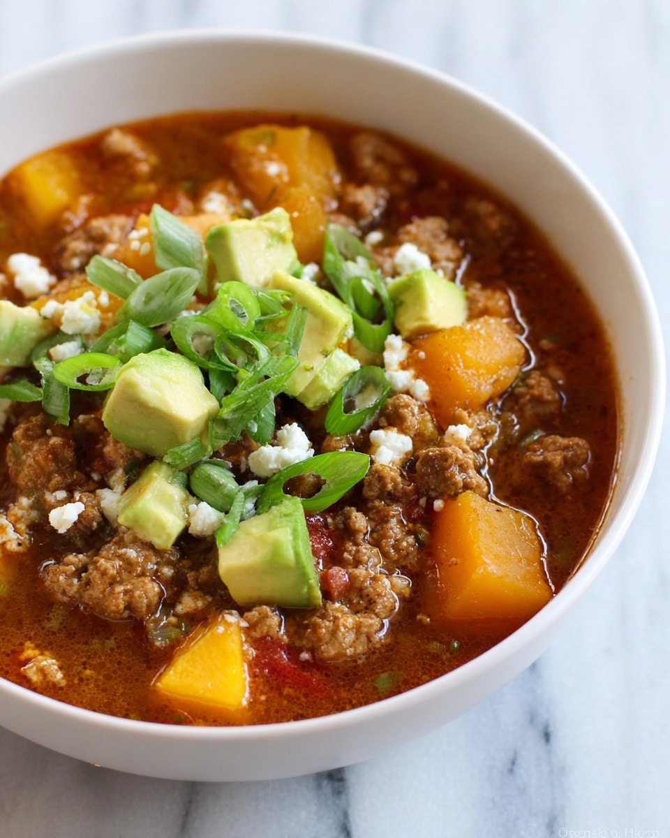 A close-up image of a bowl of chili soup with several visible layers. The base layer is a thick reddish-brown broth filled with chunks of cooked ground meat and diced yellow-orange squash. On top of this, there are scattered cubes of light green avocado and small white crumbles of cheese. Thin slices of bright green scallions are sprinkled over the whole dish, adding a fresh touch. The bowl is white and set against a white marbled surface. Photo taken with an iphone --ar 4:5 --v 7