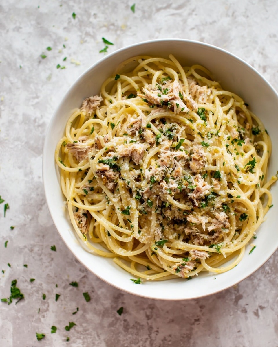 The image shows a bowl of spaghetti pasta with shredded fish mixed in, sprinkled with small green herbs and possibly grated cheese on top. The bowl is white and sits on a white marbled surface with a few scattered green herb pieces around. The pasta has a light yellow color while the fish adds a pale beige tone, and the green herbs add small pops of color. The overall look is simple and fresh, with the spaghetti noodles loosely arranged in the bowl. photo taken with an iphone --ar 4:5 --v 7