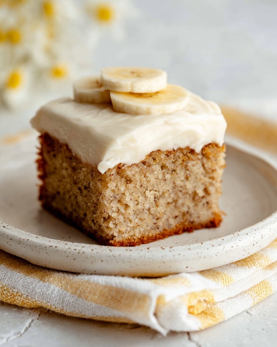 A single square piece of banana cake is placed on a small white plate with a slight speckled texture. The cake has two visible layers: the bottom layer is a moist, light brown cake with tiny dark specks, and the top layer is a creamy white frosting with a smooth and slightly thick texture. On the very top of the frosting, there is one thin round slice of banana. The plate rests on a cloth with white, yellow, and light gray stripes, and the background shows a white marbled texture. Photo taken with an iphone --ar 4:5 --v 7