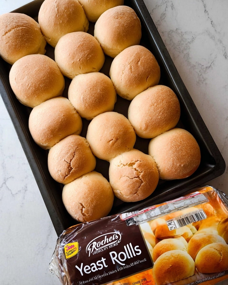 A black baking tray filled with 20 soft yeast rolls, all round and golden brown, slightly cracked on the smooth surface. The rolls are evenly placed in rows, closely packed with a warm, soft texture visible. Below the tray, there is a white and orange Rhodes bakery package labeled