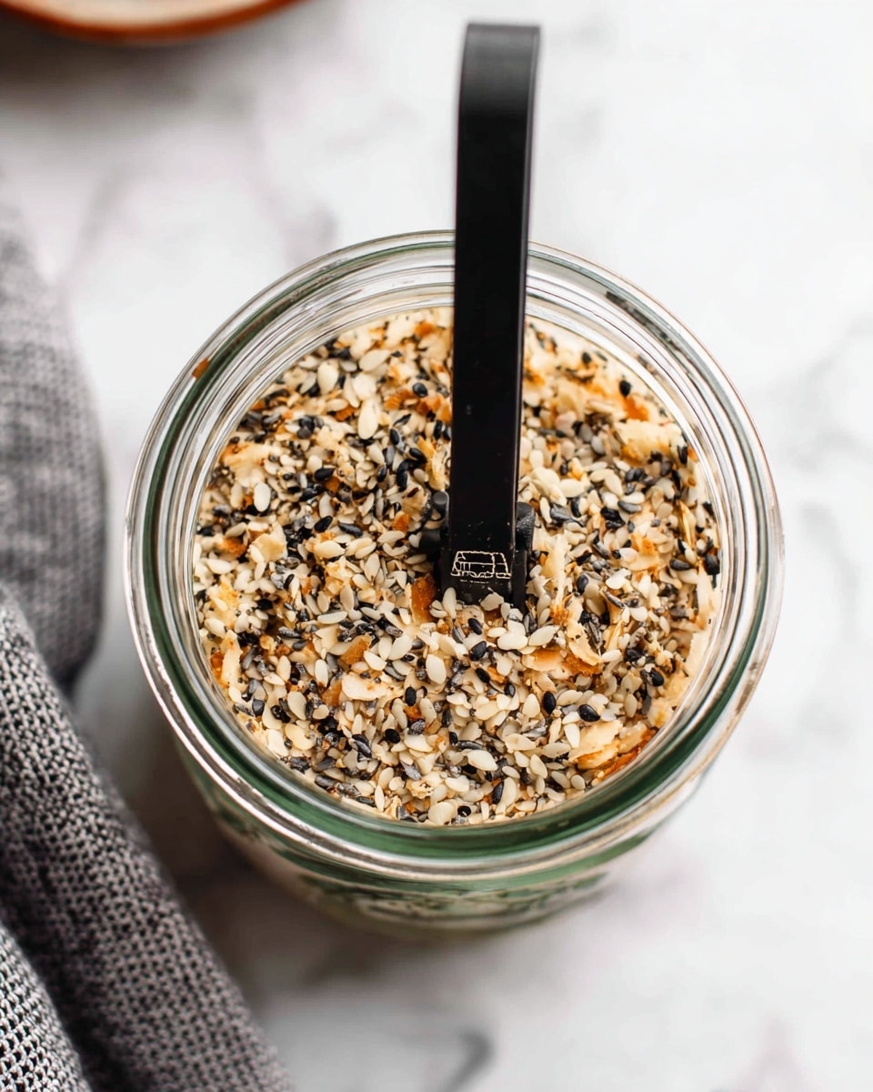 A clear glass jar filled with a textured mix of small beige, black, and white pieces resembling seeds and dried chopped onion flakes, with a pair of black measuring tongs sticking up from the jar's center. The jar is set on a white marbled surface, and a blurred grey and white checkered cloth appears on the left side of the image. Photo taken with an iphone --ar 4:5 --v 7
