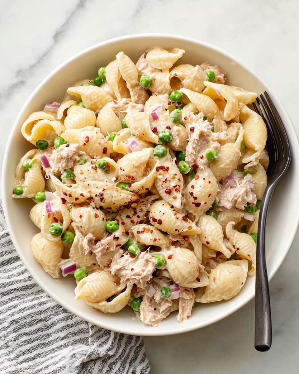 A white bowl filled with three layers of shell pasta mixed with small green peas and creamy light beige sauce. The pasta and sauce layer is scattered with small pieces of chopped purple onion, adding color contrast. The texture looks smooth and soft with hints of black pepper sprinkled on top. A silver fork rests inside the bowl on the right side. The bowl is placed on a white marbled surface with a folded white and black striped cloth napkin to the side. photo taken with an iphone --ar 4:5 --v 7