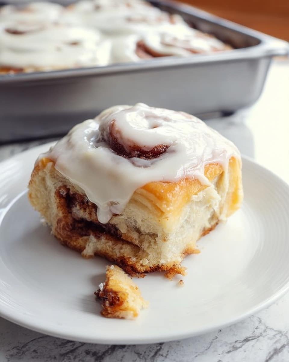 The image shows a single cinnamon roll on a white plate, placed on a white marbled texture surface. The cinnamon roll has three visible layers: a soft inner dough with a darker cinnamon swirl layer in the middle, and a thick white icing layer covering the top and slightly dripping down the sides. The roll appears fluffy with a light golden-brown color on the outer dough edges and a creamy, smooth texture of the icing. A small piece of the roll is broken off and lies next to it on the plate. In the background, there is a metal baking pan with more cinnamon rolls partially covered with white icing. Photo taken with an iphone --ar 4:5 --v 7