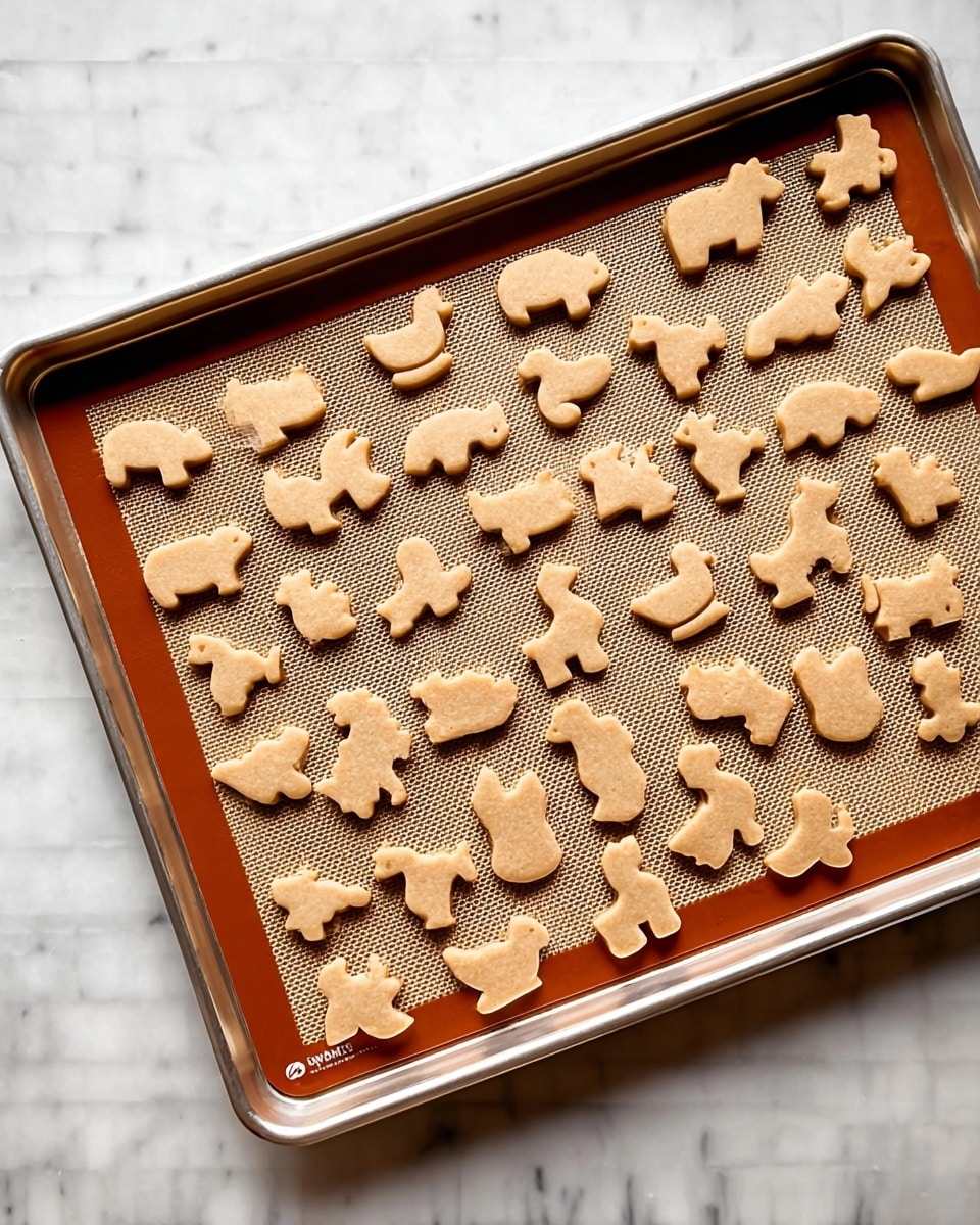 A metal baking tray holds about fifty small animal-shaped cookies arranged neatly over a tan silicone baking mat with a slight grid pattern. The cookies have a light brown color and simple, smooth surfaces, shaped like various animals including bears, ducks, rabbits, horses, and sheep, spread evenly across the tray. The tray sits on a white marbled surface, with the warm color of the cookies and mat contrasting against the clean, bright background. photo taken with an iphone --ar 4:5 --v 7