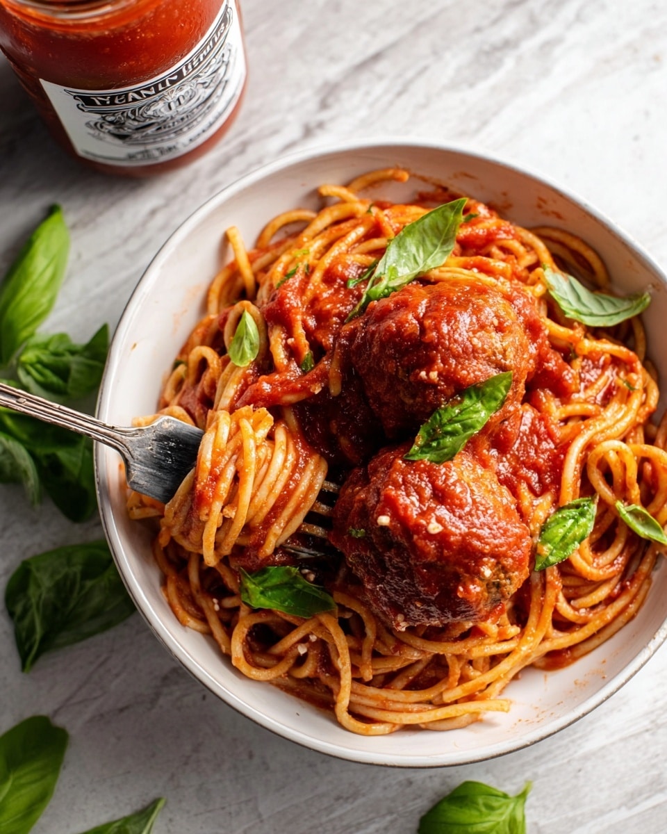 A white bowl filled with two large, round meatballs covered in rich red tomato sauce sitting on a bed of thick spaghetti noodles also coated in sauce. The spaghetti is twisted around a silver fork that dips into the bowl. Fresh green basil leaves are scattered on top and around the dish, adding a pop of color. The bowl is placed on a white marbled surface with a jar of tomato sauce behind it in the background. photo taken with an iphone --ar 4:5 --v 7