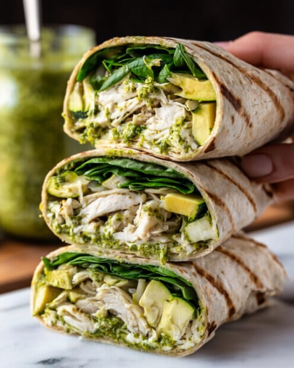 The image shows a stack of three wrapped sandwiches with the top one cut in half to reveal the inside. Each wrap is light brown with grill marks on the outside. Inside, there are distinct layers: at the bottom, fresh green spinach leaves; above that, slices of yellow grilled zucchini; next, shredded white chicken pieces; and on the very top, a bright green spread that looks like pesto. The stack rests on a white marbled surface, with a jar of green sauce slightly blurred in the background. A woman's hand is gently holding the top sandwich half. Photo taken with an iphone --ar 4:5 --v 7