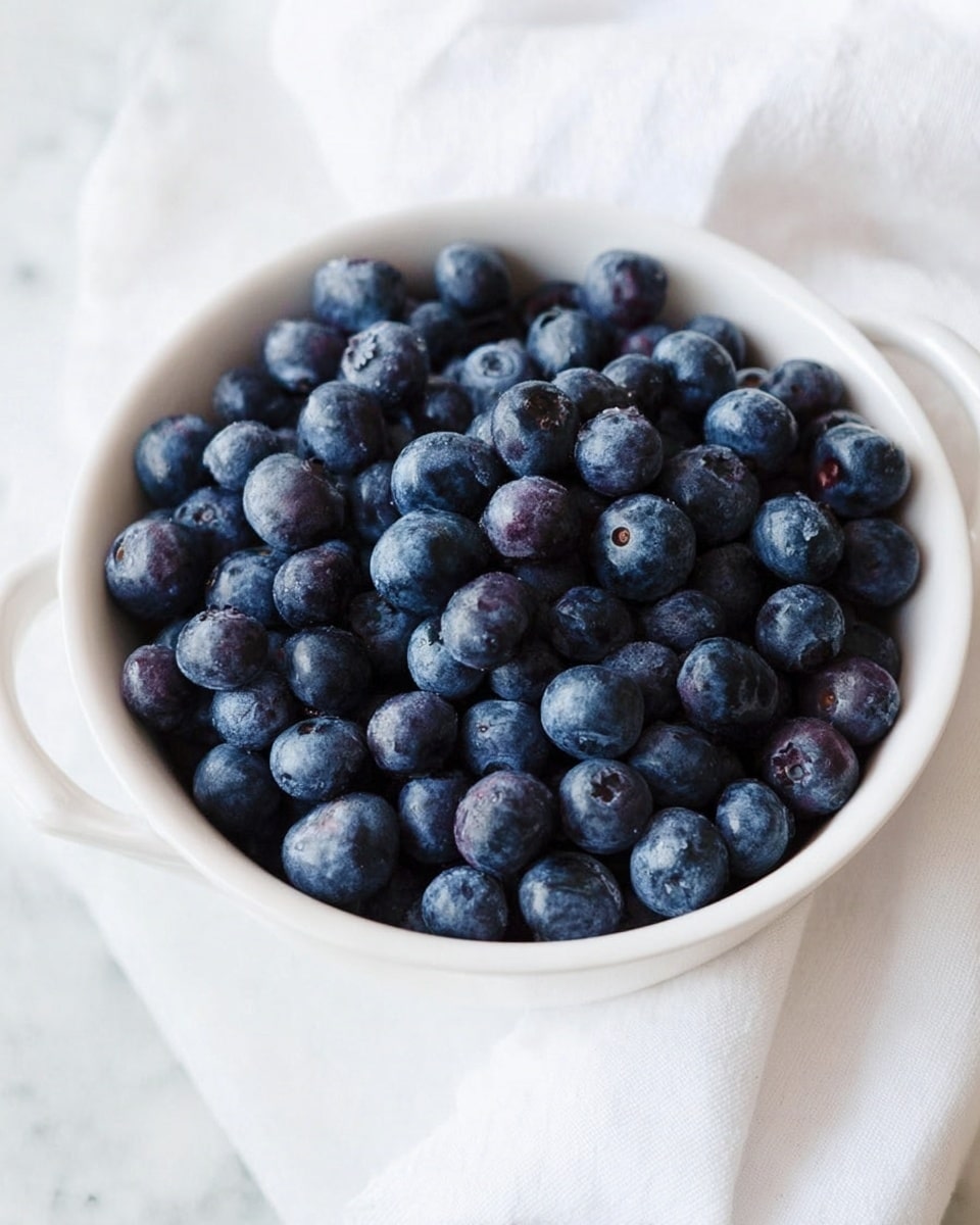 A white bowl filled to the top with fresh blueberries, showing their round shape and deep blue to purple color with a slight powdery shine on the skin. The bowl sits on a white marbled surface with a white cloth partially visible beside it, and the scene is brightly lit with soft natural light that highlights the texture of the berries. photo taken with an iphone --ar 4:5 --v 7