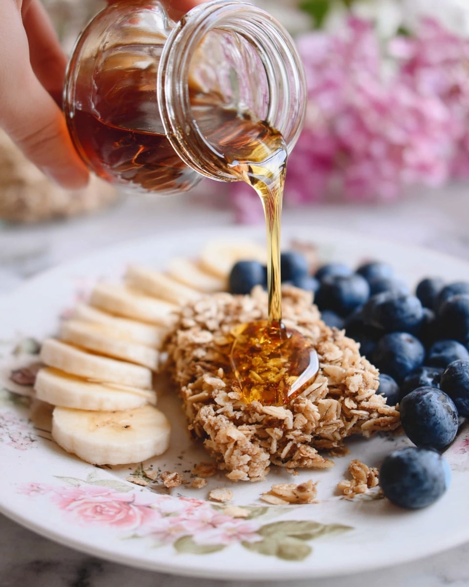 A close-up image shows a glass jar pouring golden syrup over a granola bar on a white plate with soft floral patterns on the edges. The granola bar has a rough texture with visible oats pressed together in a rough rectangular shape. Around the granola bar, fresh and plump blueberries are placed on the left side, next to slices of banana arranged in a neat line. The background features a white marbled surface with some blurred pink flowers in the distance. A woman's hand is holding the jar from above. photo taken with an iphone --ar 4:5 --v 7