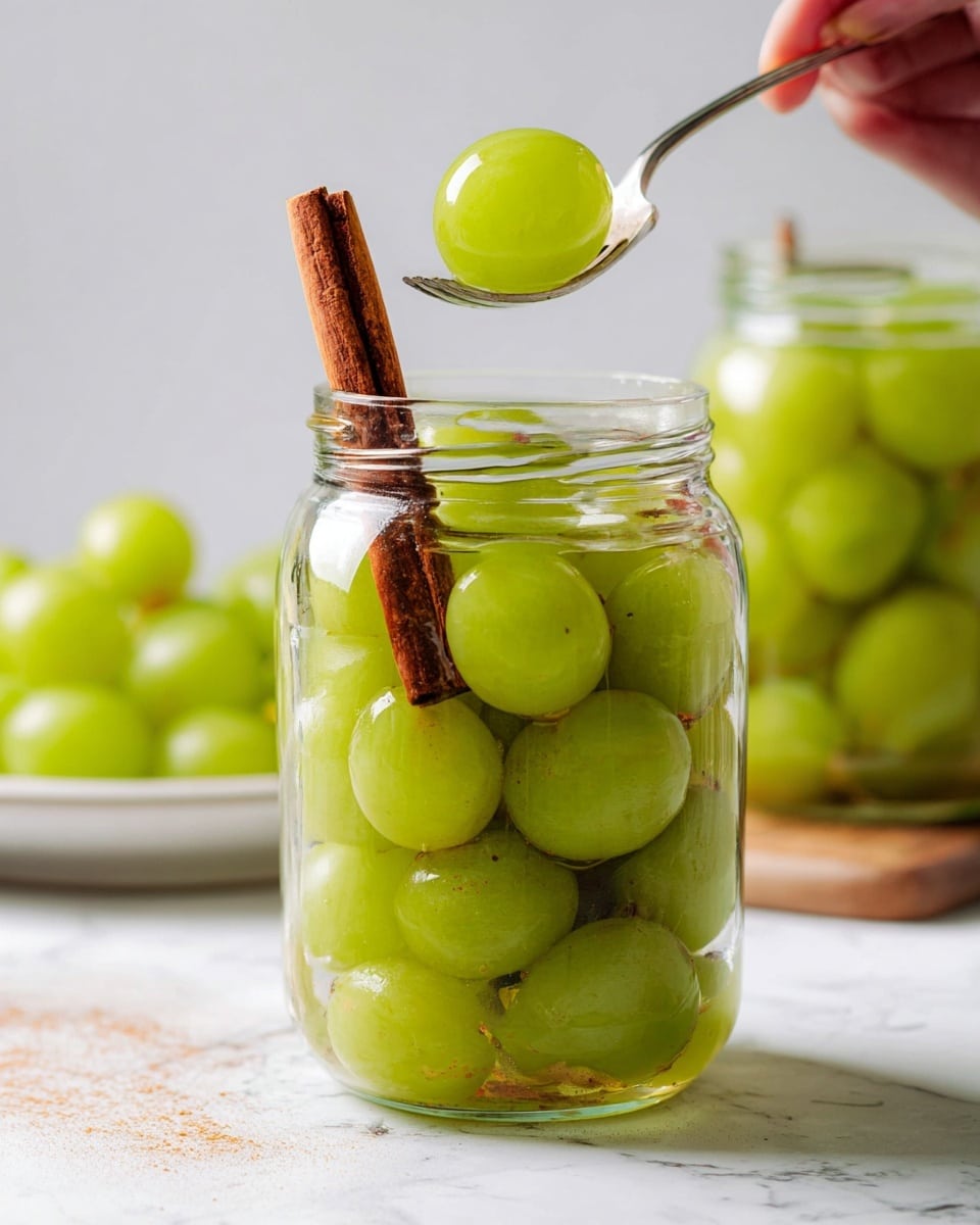 A clear glass jar filled with bright green grapes soaked in a light liquid, with a single brown cinnamon stick inside the jar leaning diagonally. A silver fork lifted by a woman's hand is holding one shiny green grape above the jar’s open mouth. Behind the jar, another glass jar filled similarly is partially visible. On the left side, a white plate holds more green grapes. The scene is set on a white marbled surface with two cinnamon sticks lying down near the jar. The light reflects softly on the glass and grapes, showing freshness and clarity photo taken with an iphone --ar 4:5 --v 7