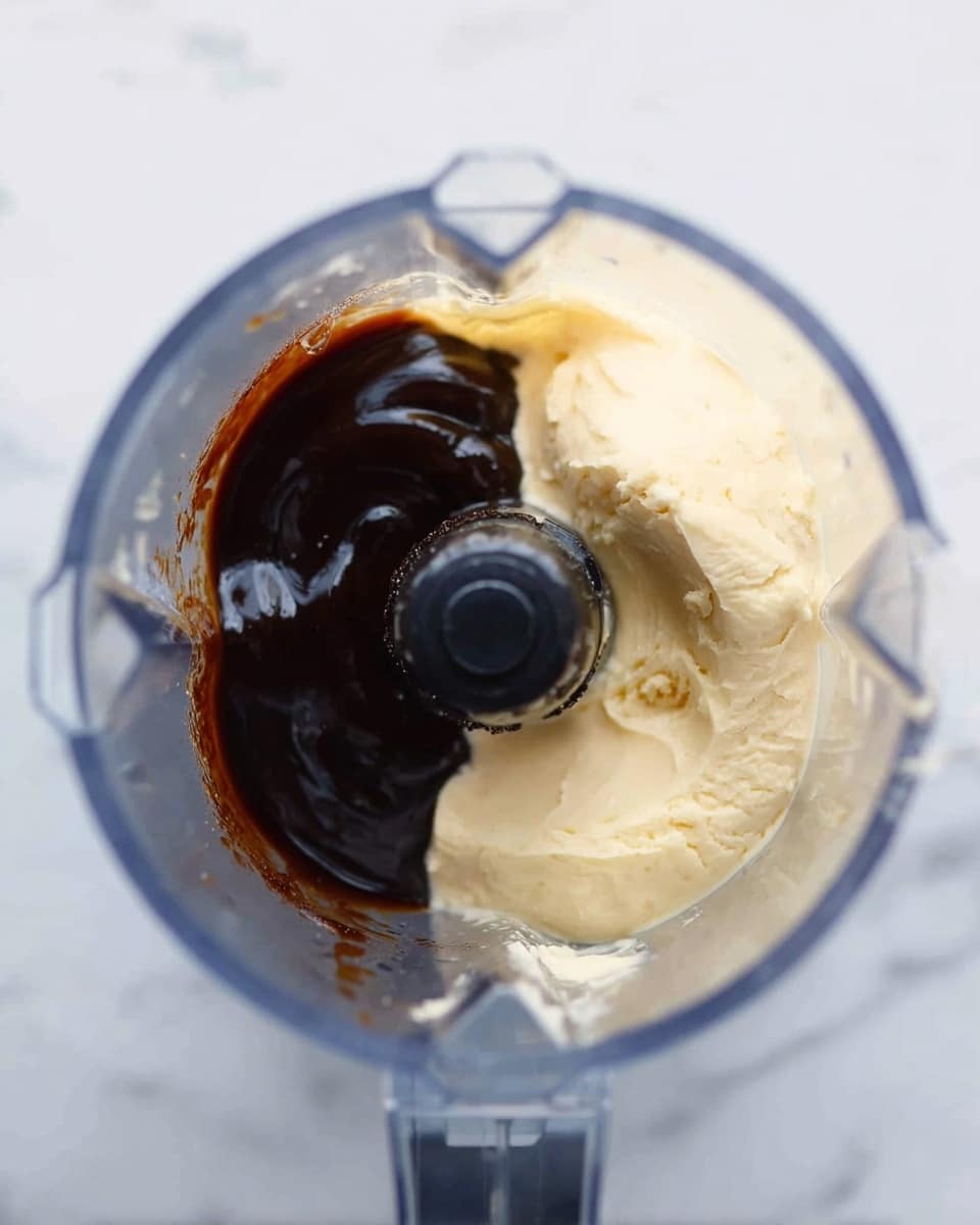 The image shows a close-up of a blender with two main layers inside: on the right side, there is a thick pale yellow creamy substance with a smooth and slightly rough texture, and on the left side, there is a dark brown sauce with a shiny, smooth, and glossy texture. The blender blades are visible in the middle, surrounded by the two layers. The background has a white marbled texture. Photo taken with an iphone --ar 4:5 --v 7