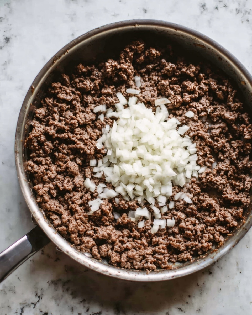 A silver pan filled with cooked brown ground meat evenly spread across the bottom, topped in the center with a small pile of white chopped onions. The pan is placed on a surface with a white marbled texture. Photo taken with an iphone --ar 4:5 --v 7
