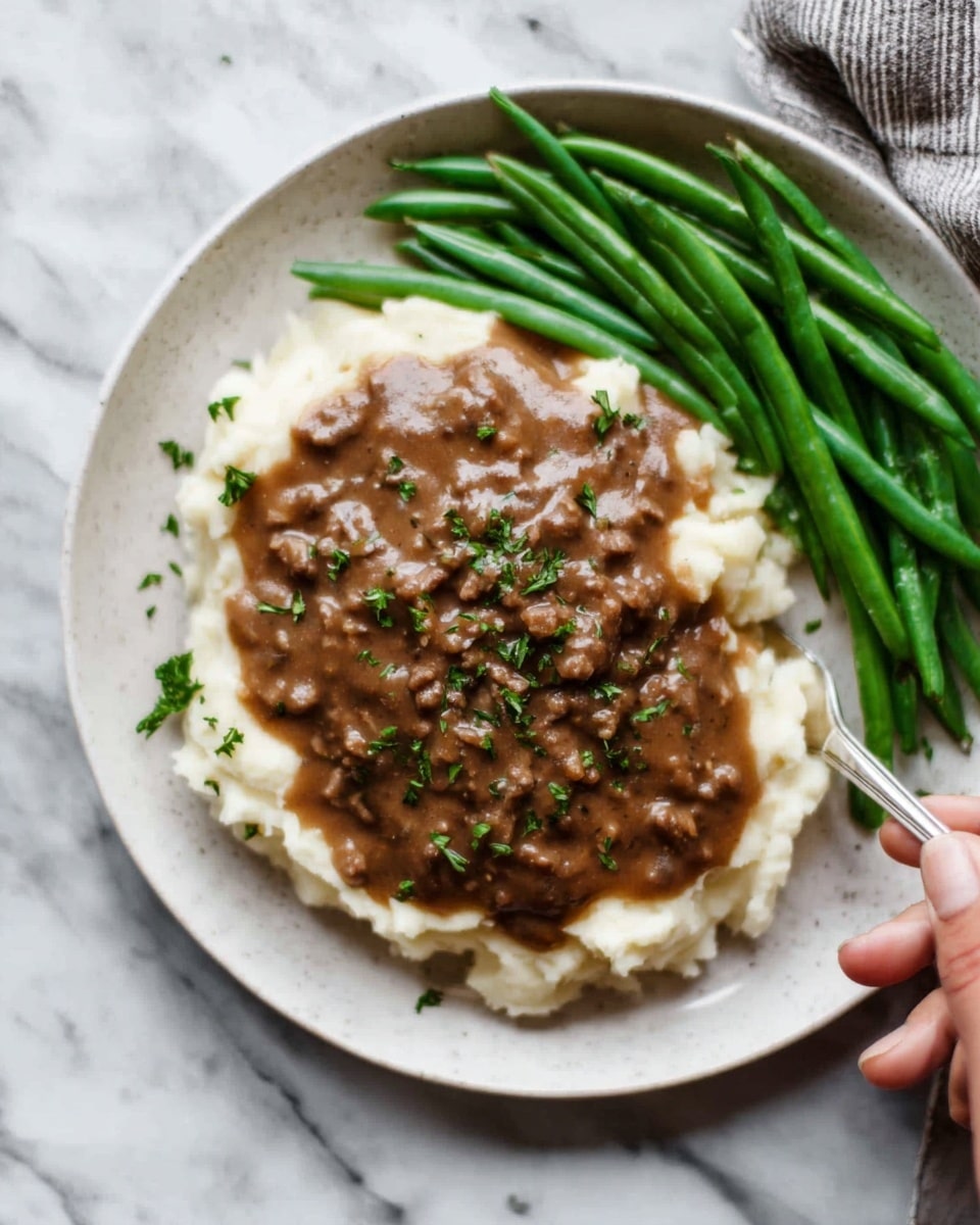 A white plate holds a layer of smooth, fluffy mashed potatoes as the base, topped with a rich, chunky brown gravy mixed with small bits of meat spread evenly over the center. On the right side, there is a neat pile of vibrant green beans with a slight shine, arranged in a straight line. The dish is sprinkled with finely chopped green herbs on top of the gravy. The plate sits on a white marbled surface, and a woman's hand holding a fork is reaching towards the plate. photo taken with an iphone --ar 4:5 --v 7