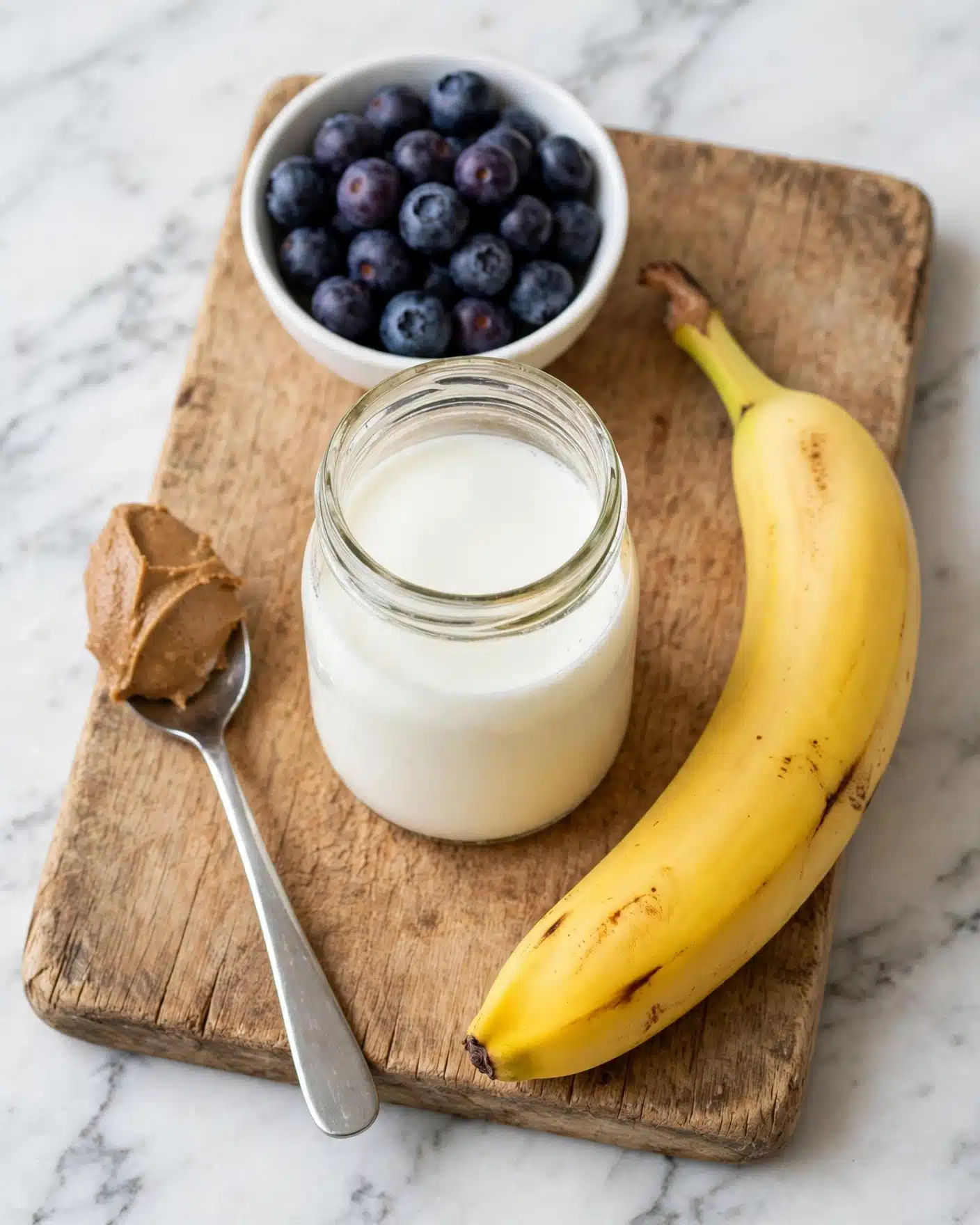 The image shows a wooden cutting board with four items placed on it. There is a small white bowl at the top filled with dark purple blueberries, a glass jar in the middle filled with white milk, a bright yellow banana positioned diagonally on the right side, and a silver spoon on the left holding a scoop of light brown almond butter. The background is a white marbled surface. Photo taken with an iphone --ar 4:5 --v 7