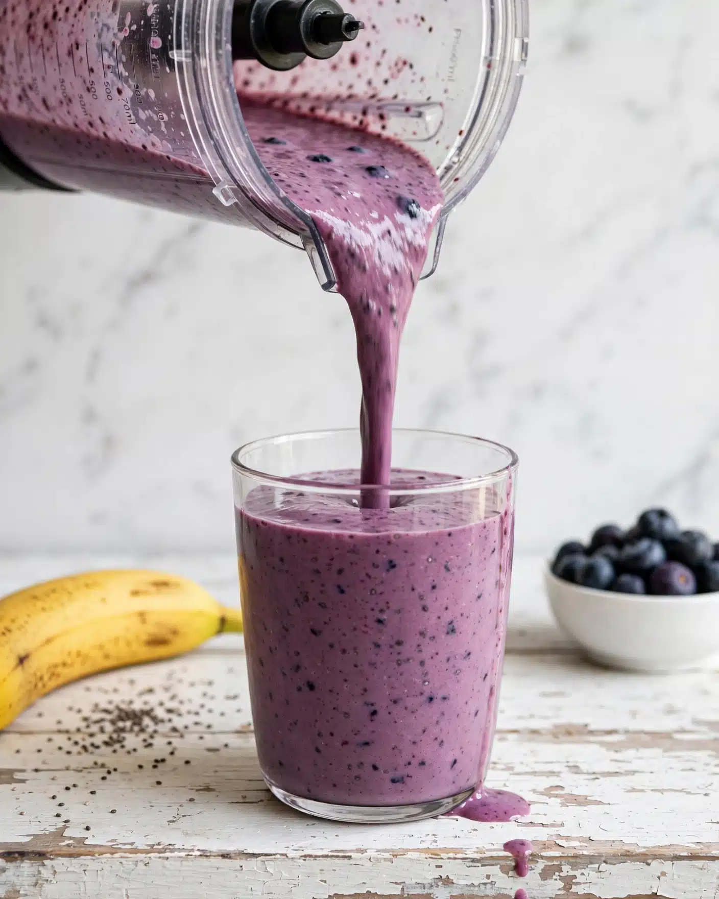 A thick purple smoothie is being poured from a clear blender container into a clear glass, nearly filling it with a smooth, creamy texture and small dark flecks throughout. The glass sits on a rustic, white distressed wooden surface with a drop of smoothie spilled on the edge. In the background, there is a yellow banana lying on the left side, a small white bowl filled with dark blueberries on the right, and a small amount of chia seeds scattered near the banana. The background is a white marbled texture. Photo taken with an iphone --ar 4:5 --v 7