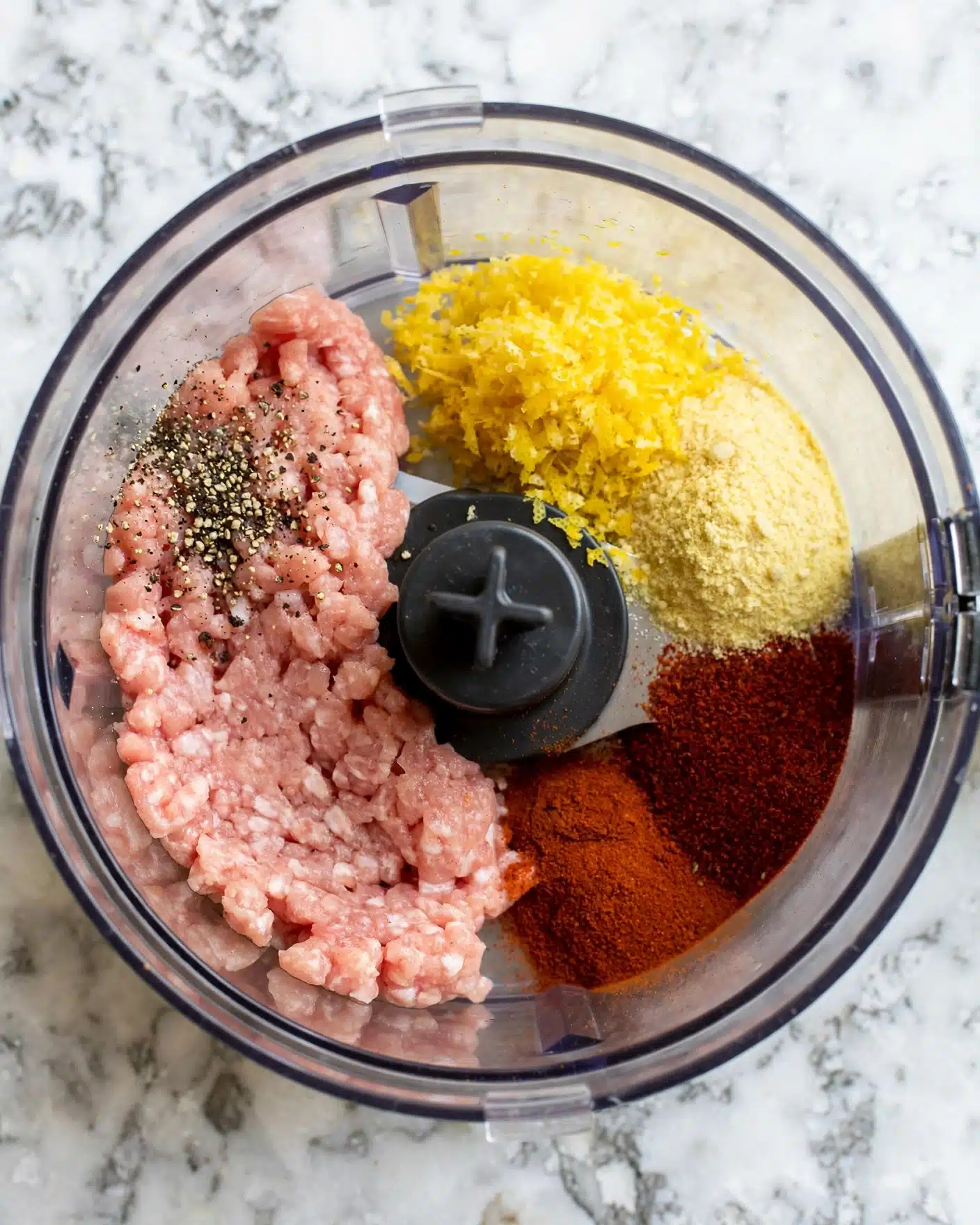 Homemade Chicken Nuggets Recipe 4 A top view of a food processor bowl filled with several ingredients arranged in separate sections. There is a pale pink layer of raw meat on the left side, surrounded by dark black and white pepper specks on the upper left, a rough yellow lemon zest and a light yellow powder on the upper right, and at the bottom right, a bright reddish-brown spice and a dark reddish spice. The food processor has a black center blade in the middle, and the whole image is set on a white marbled texture photo taken with an iphone --ar 4:5 --v 7