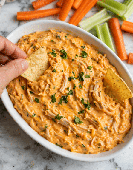 A white oval dish filled with a thick, creamy orange dip that has visible strands of shredded chicken mixed throughout, topped with small bits of chopped green herbs. Two light yellow corn chips are dipped into the dip, one held by a woman's hand and the other resting partially inside. In the background, there are fresh carrot sticks and celery pieces on a white marbled surface. The dip looks warm and slightly textured, with a rich and cheesy appearance photo taken with an iphone --ar 4:5 --v 7