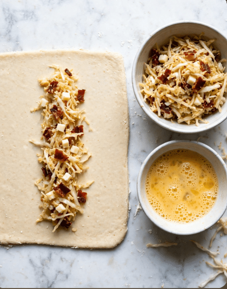 A square sheet of light beige dough is placed on a white marbled surface, with a narrow strip of filling in the center made of shredded light tan cabbage, small white cheese cubes, and small dark red pieces resembling dried meat or bacon. To the right, there's a white bowl filled with more of the same shredded cabbage mix with cheese and dried meat pieces, and below that, another white bowl contains a beaten yellow egg mixture with bubbles on the surface. The scene shows a neat preparation for wrapping or folding the dough. Photo taken with an iphone --ar 4:5 --v 7