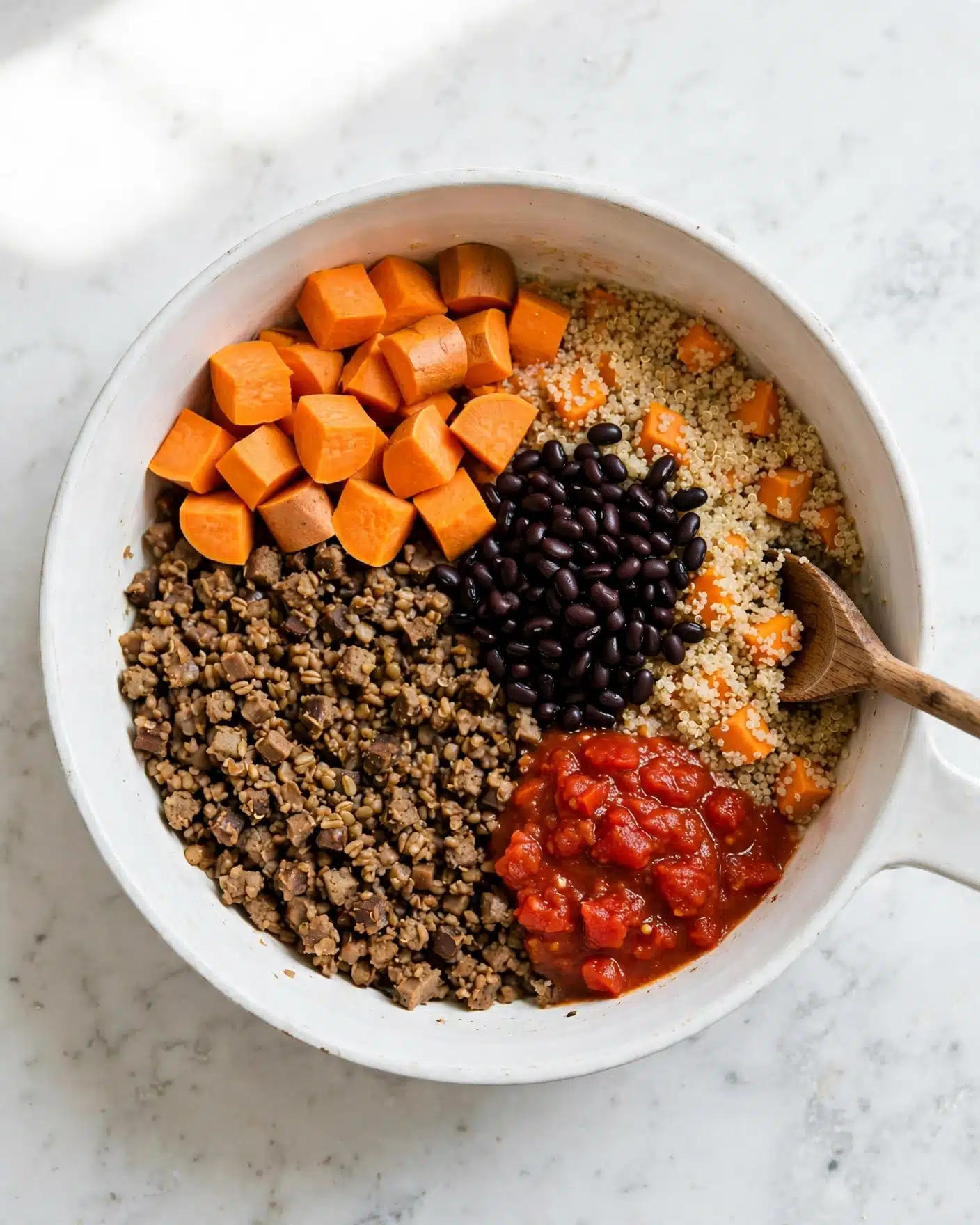 Black Bean, Sweet Potato, and Quinoa Chili with Smoked Chipotle Recipe 4 A large white pot contains four main visible layers. In the bottom left and center, there are small cooked brown pieces, likely vegetables or grains, forming the base layer with a slightly textured look. On the top left, there are bright orange chunks, which seem to be raw or lightly cooked sweet potatoes, grouped together in a neat pile. To the right, there is a layer of light beige quinoa mixed with some orange chunks. On top of the quinoa and next to the sweet potatoes is a pile of shiny black beans. There is also a bright red sauce with a chunky texture near the bottom right of the pot. A wooden spoon rests on the right side inside the pot, touching the quinoa. The whole pot sits on a white marbled surface. photo taken with an iphone --ar 4:5 --v 7