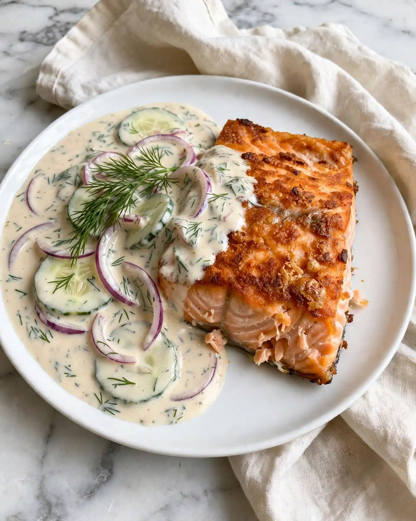 Grilled Salmon with Creamy Cucumber-Dill Salad Recipe 5 A piece of cooked salmon with a crispy orange-brown top layer, placed on a white plate, slightly to the right. On the left side of the salmon, there is a creamy sauce with visible slices of cucumber and red onion mixed with dill herbs, covering part of the fish and spreading towards the plate's edge. A sprig of fresh dill rests on top of the creamy sauce, adding green color and texture. The plate sits on a folded light-colored cloth on a white marbled surface. Photo taken with an iphone --ar 4:5 --v 7