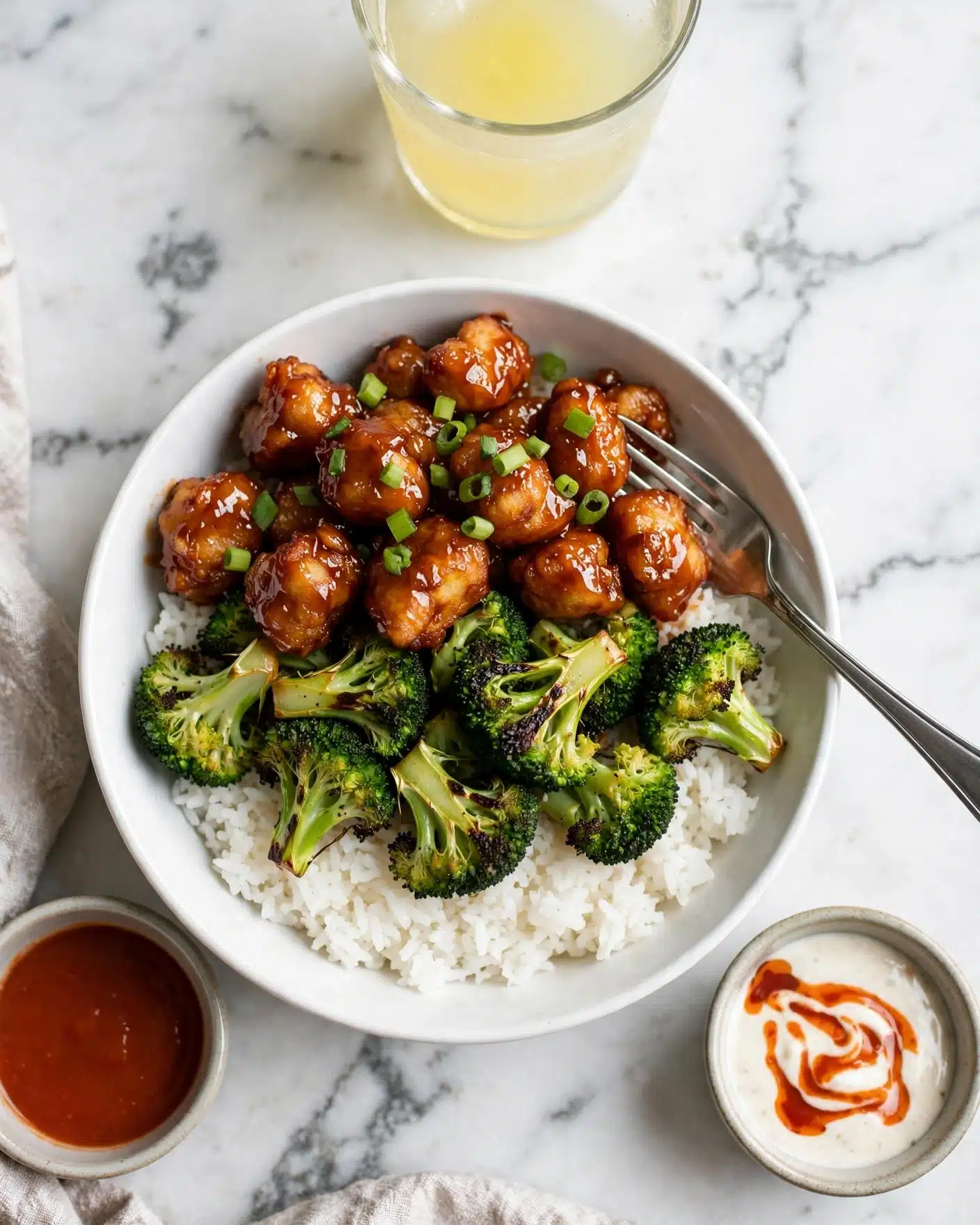 Sheet Pan Honey Buffalo Chicken with Broccoli Recipe 5 A white bowl filled with three layers: the bottom layer is fluffy white rice, the middle layer is bright green roasted broccoli florets with some charred edges, and the top layer is golden-brown fried pieces coated in a shiny reddish-brown sauce, garnished with small chopped green onions. A silver fork is placed on the right side of the bowl, slightly touching the food. The bowl is placed on a white marbled surface, with a glass of light yellow liquid above it and small containers of reddish sauce and white creamy sauce with reddish drizzle nearby. Photo taken with an iphone --ar 4:5 --v 7