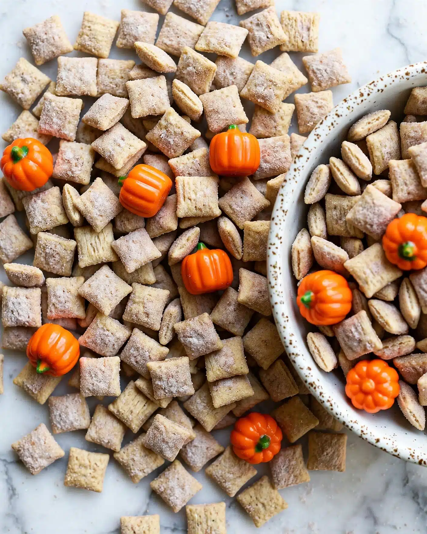 Pumpkin Spice Puppy Chow Recipe 5 The image shows a close-up of a pile of small, square cereal pieces covered lightly with a powdered cinnamon sugar mix, giving them a soft beige color with a slightly powdery texture. Scattered among the cereal are bright orange candy pumpkins with smooth, shiny surfaces and small green stems, adding a vibrant contrast to the warm tones of the cereal. On the right side, a white bowl with a speckled brown rim is partially visible, half-filled with more of the cereal and candy pumpkins spilling out onto a white marbled surface. photo taken with an iphone --ar 4:5 --v 7