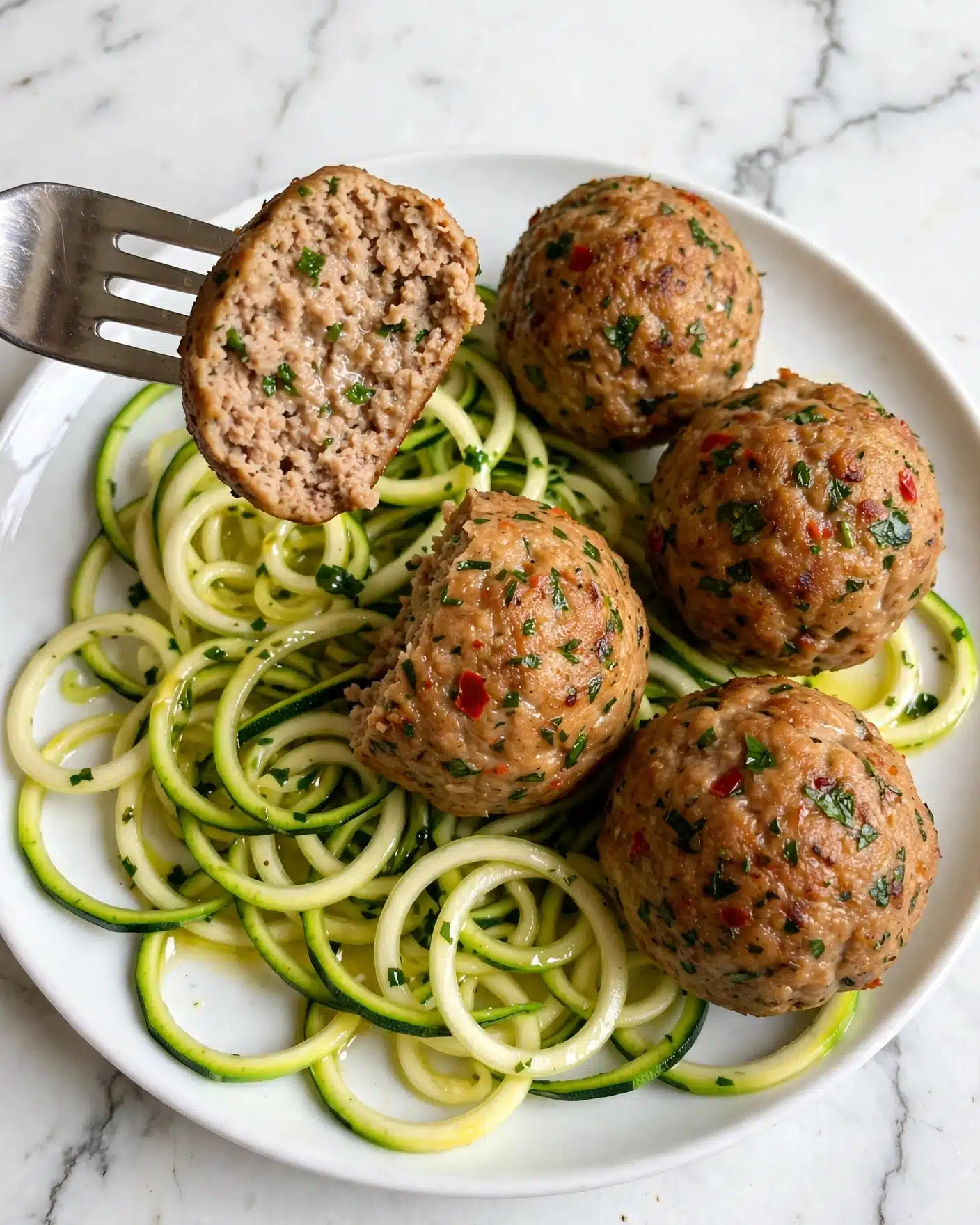 Easy Baked Chicken Meatballs with Lemon Herb Butter Sauce Recipe 5 The image shows five golden-brown meatballs that have small green and red herb bits on their surface, giving them a textured, seasoned look. One meatball is cut open, showing a light, moist inside with specks of green herbs, held by a silver fork. Below the meatballs, there is a bed of thin, spiral-shaped zucchini noodles that are light green and yellow, fresh and slightly shiny. The whole set of food sits on a white plate placed on a white marbled surface. The photo taken with an iphone --ar 4:5 --v 7