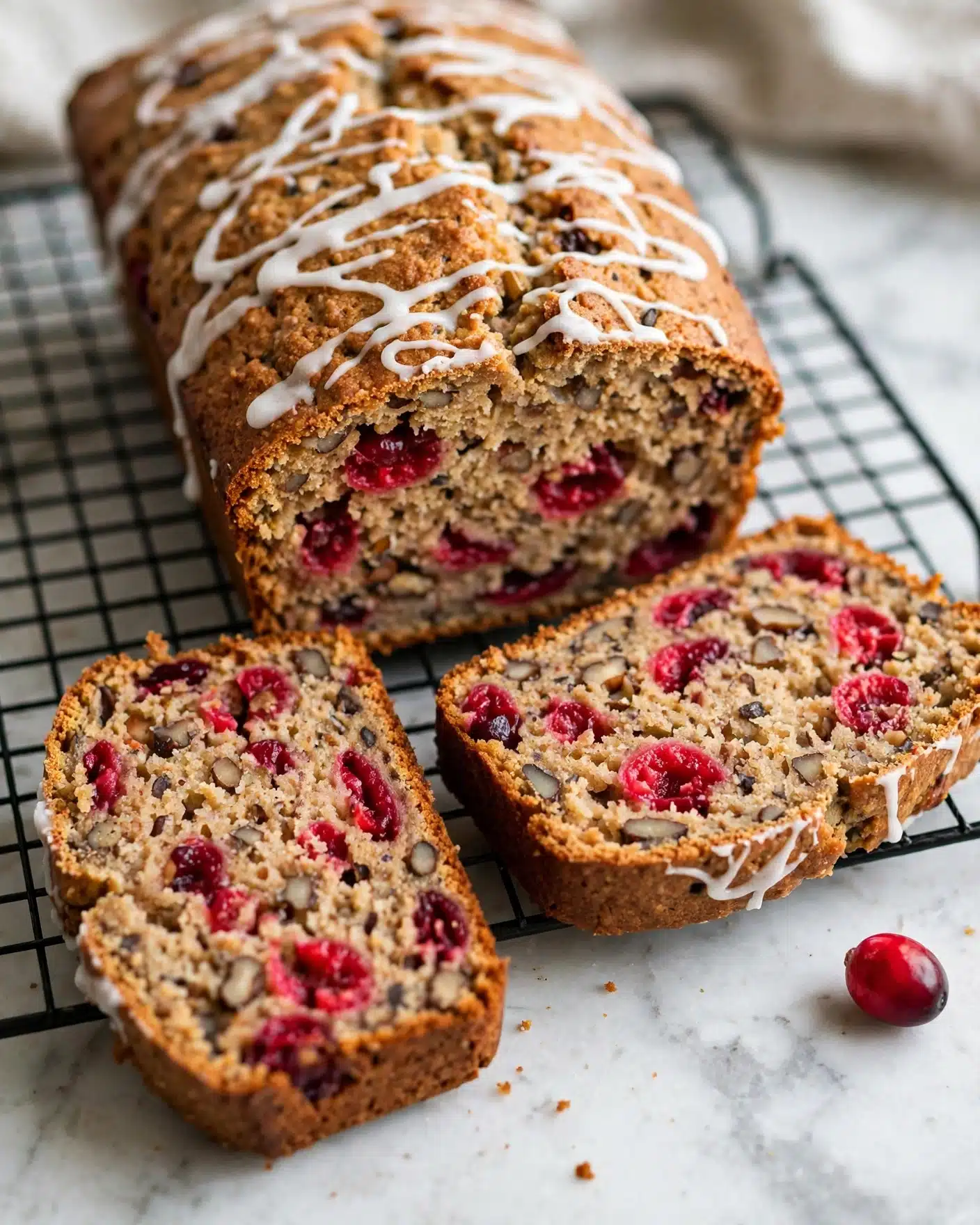 Orange Glazed Cranberry Bread Recipe 5 A loaf of cranberry nut bread is shown on a black wire cooling rack with two slices cut and laid in front of the main loaf. The bread is golden brown with a slightly rough texture, filled with bright red cranberries and small dark nuts scattered throughout. The top has a light drizzle of white icing in a loose pattern, contrasting with the golden crust. A single fresh cranberry lies on the white marbled surface nearby. The close-up view shows the moist, dense inside texture, highlighting the fruit and nut pieces. Photo taken with an iphone --ar 4:5 --v 7