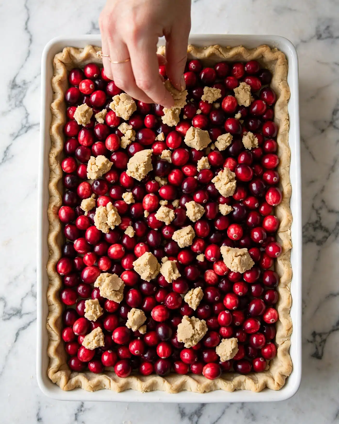Cranberry Shortbread Bars Recipe 4 A rectangular white pie dish contains three visible layers. The bottom layer is a light beige pie crust pressed evenly into the dish with slightly crimped edges. The middle layer is a thick spread of red and dark red fresh cranberries scattered evenly across the crust, covering it fully. The top layer is uneven clusters of light tan dough being placed by a woman's hand with a gold ring, gently pinching a piece of the crumbly dough above the cranberries. The dish is set on a white marbled surface with soft natural light highlighting the colors and textures. Photo taken with an iphone --ar 4:5 --v 7