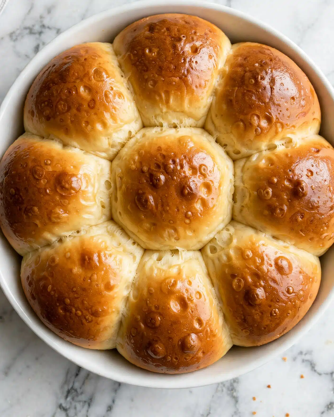 Texas Roadhouse Rolls with Cinnamon Honey Butter Recipe 5 The image shows a close-up of a white dish filled with softly baked golden brown rolls. There are multiple rolls lined up in rows, each roll having a smooth, shiny top with a slightly lighter, soft-looking side that touches the other rolls. The tops have a warm golden color with some areas darker than others, showing a baked texture. The surface of the rolls is smooth but with small air bubbles visible in places, indicating light fluffiness inside. The dish sits on a white marbled texture background photo taken with an iphone --ar 4:5 --v 7