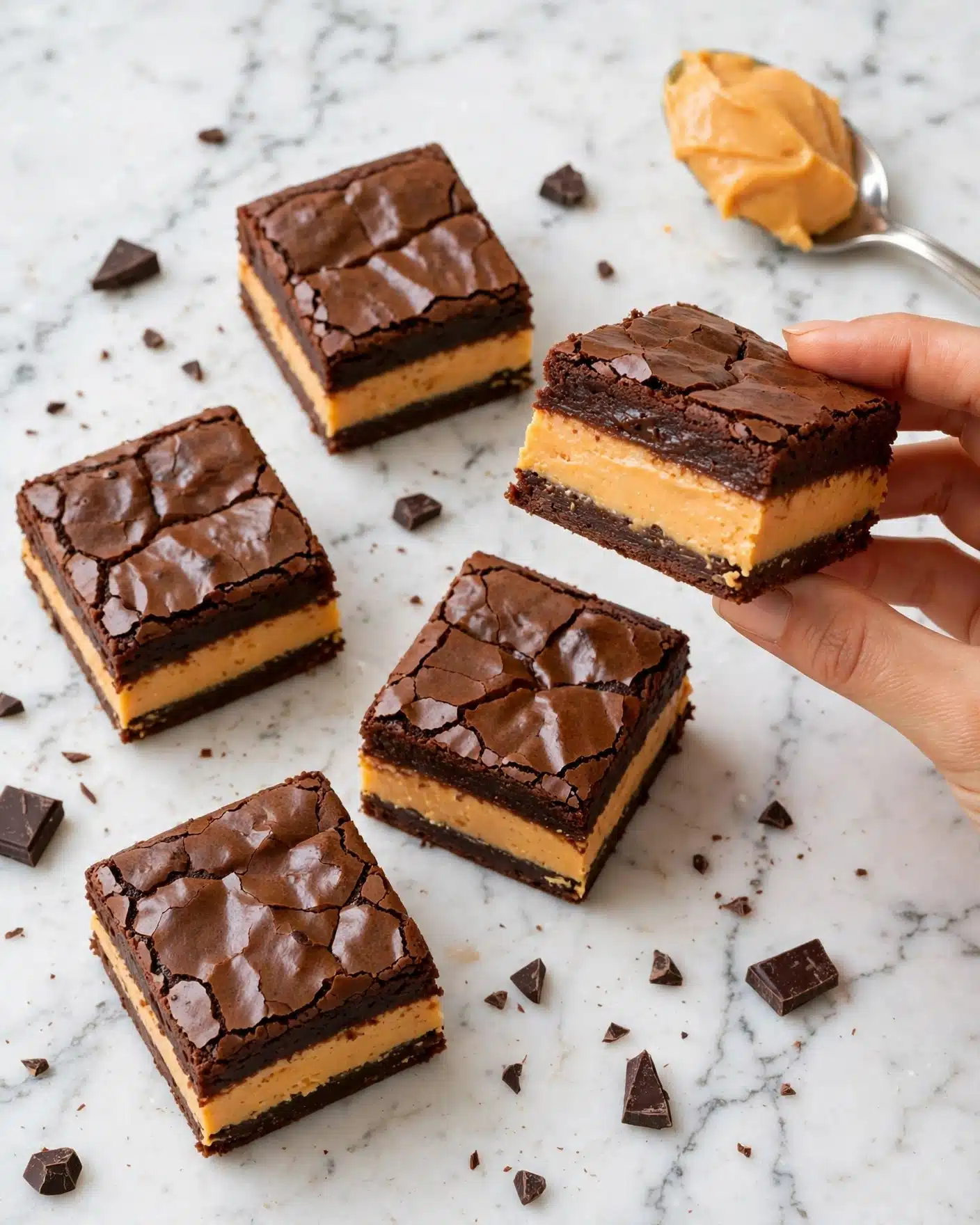 Pumpkin Cream Cheese Swirl Brownies Recipe 5 The image shows several square brownie pieces placed on a white marbled surface. Each brownie has two clear layers: a dark brown, shiny, slightly cracked top layer with a smooth chocolate texture, and a thick, dense, light brown middle layer that looks like peanut butter or caramel. Small chunks of chocolate scatter around the brownies. A silver spoon with a dollop of orange creamy filling rests near the top right corner. One brownie is held up in the air by a woman's hand, showing the side view of the two contrasting layers clearly. photo taken with an iphone --ar 4:5 --v 7